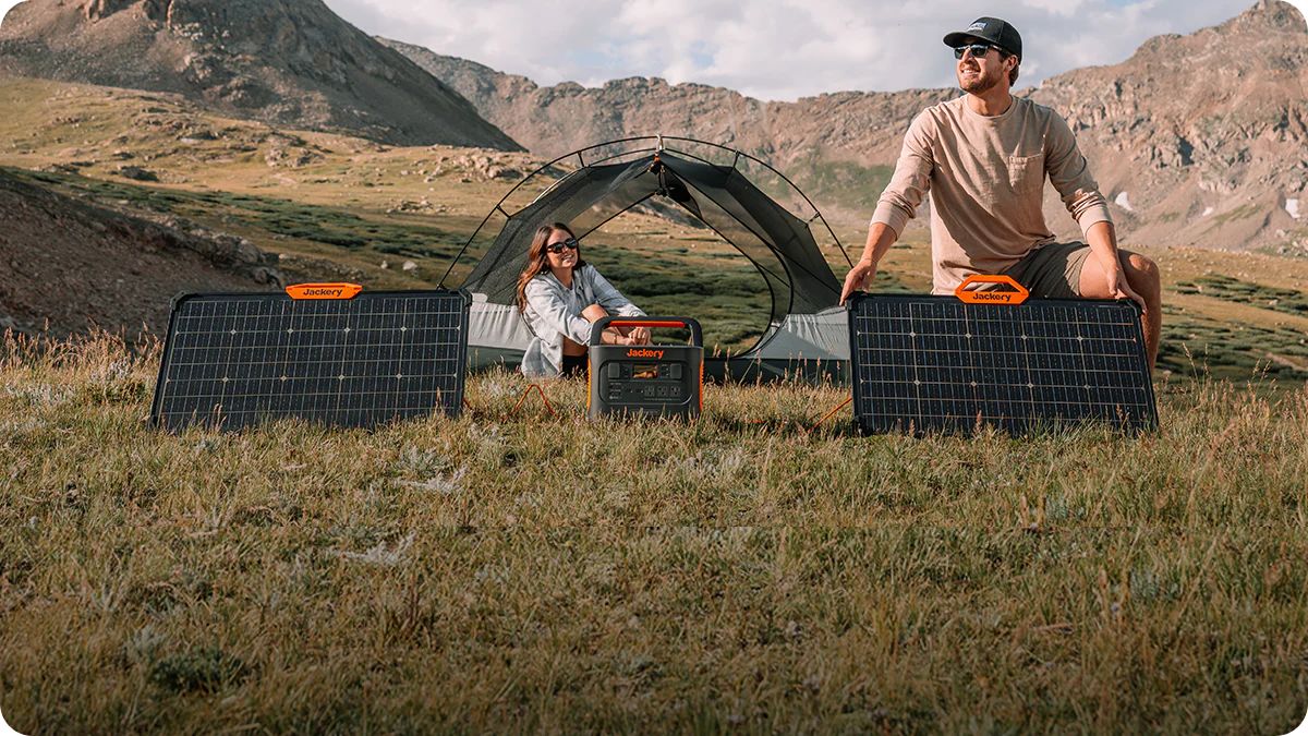 Twee kampeerders in de bergen bij een tent met een draagbare powerstation, aangesloten op twee draagbare zonnepanelen die in het gras staan opgesteld.