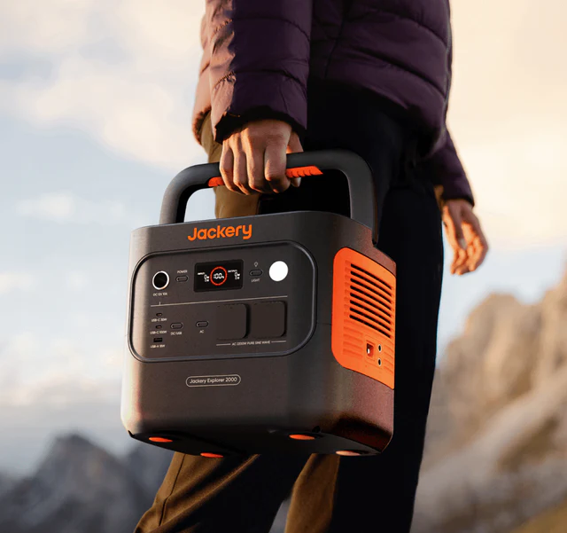 Jackery Explorer 2000 draagbaar powerstation zwart met oranje accenten, in hand van persoon buiten met berglandschap op de achtergrond