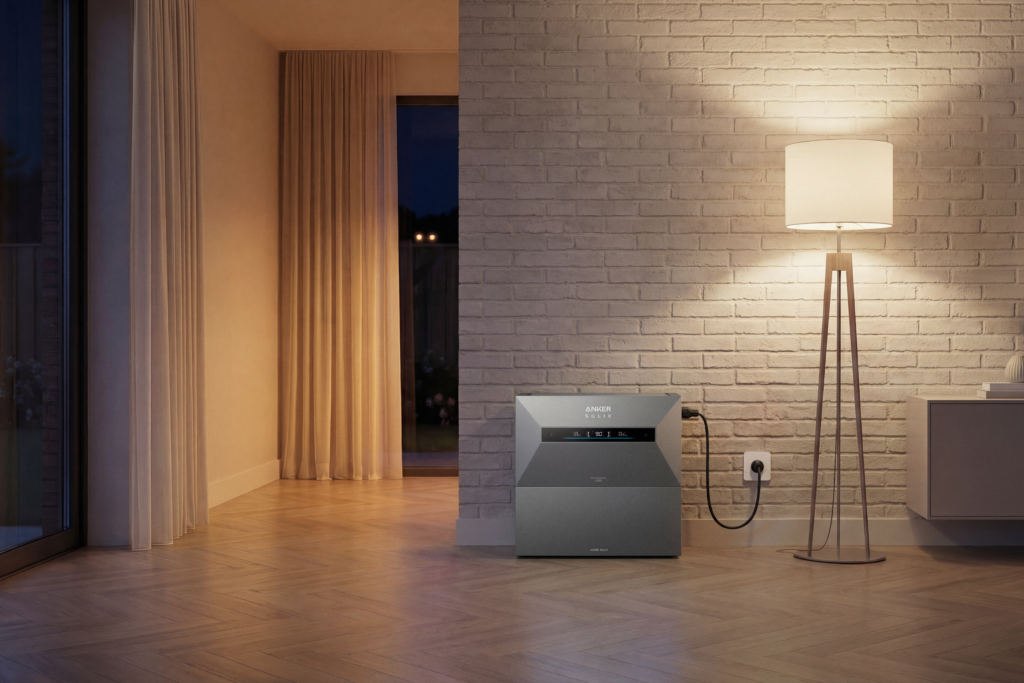 Anker Solix grijze thuisbatterij geplaatst tegen een wit bakstenen muur in een woonkamer, aangesloten op een wandcontactdoos, naast een moderne houten vloerlamp met witte lampenkap.