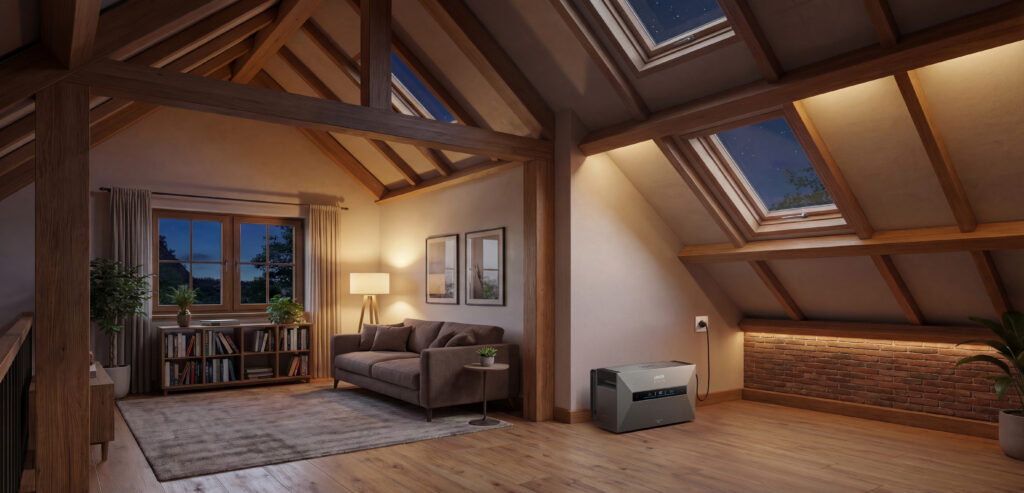 Portable powerstation Anker, gebruikt als kleine thuisbatterij, met grijze behuizing in een sfeervolle zolderkamer met houten balken, dakramen en warme verlichting naast een bakstenen muur, aangesloten op het stopcontact voor off-grid energievoorziening.